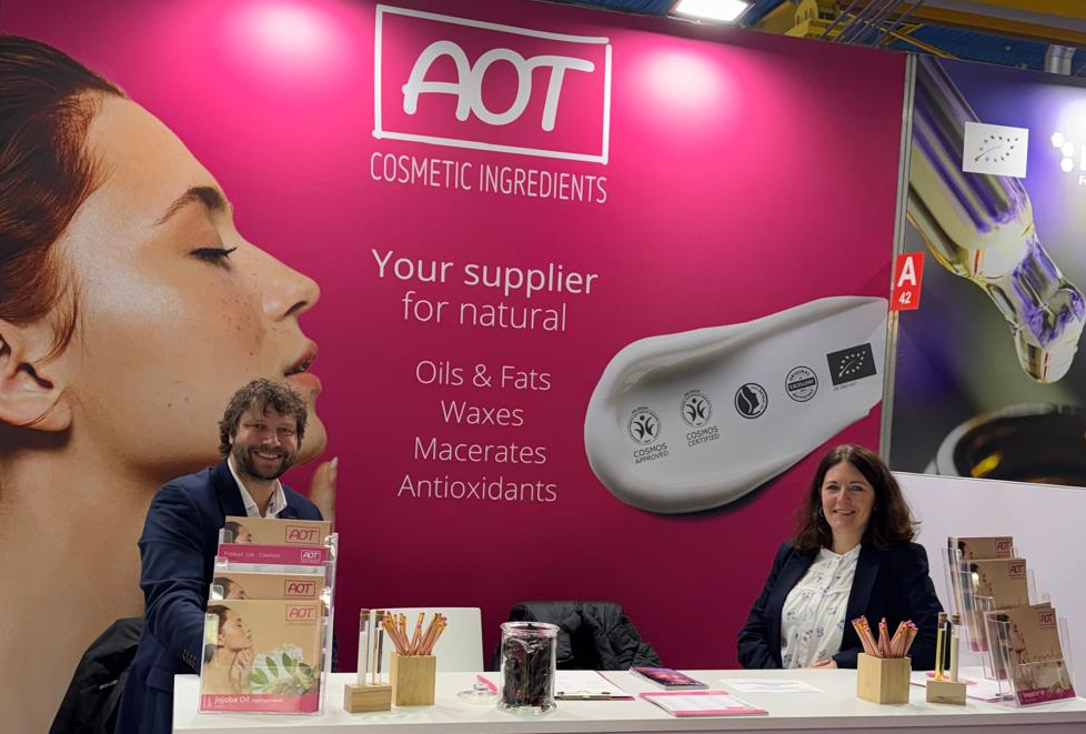 AOT trade fair stand at Cosmoprof 2026 with the team in front of a pink back wall reading “Your supplier for natural ingredients”